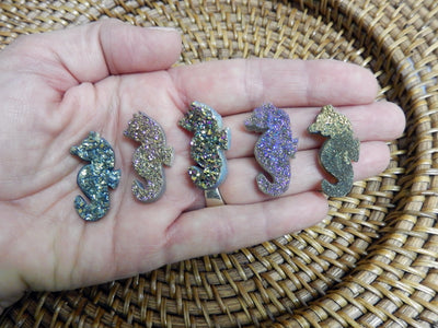 Hand holding up 5 Rainbow Titanium Treated Druzy Seahorse Shaped Cabochons in front of woven basket