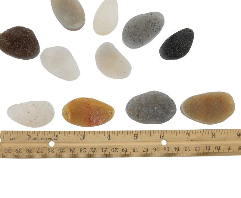 12 assorted druzy cabachons to show they vary in color from white, gray, black, brown, tan, orange, purple.  They have a drill hole on each side. Shown with a ruler to show they are about an inch and a half but will vary.