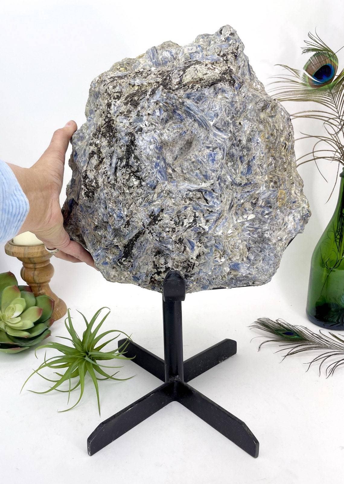hand next to Large Blue Kyanite on Metal Stand with decorations in the background