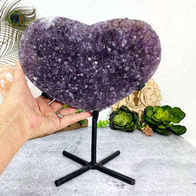 hand next to Amethyst Druzy Heart On Metal Stand with decorations in the background