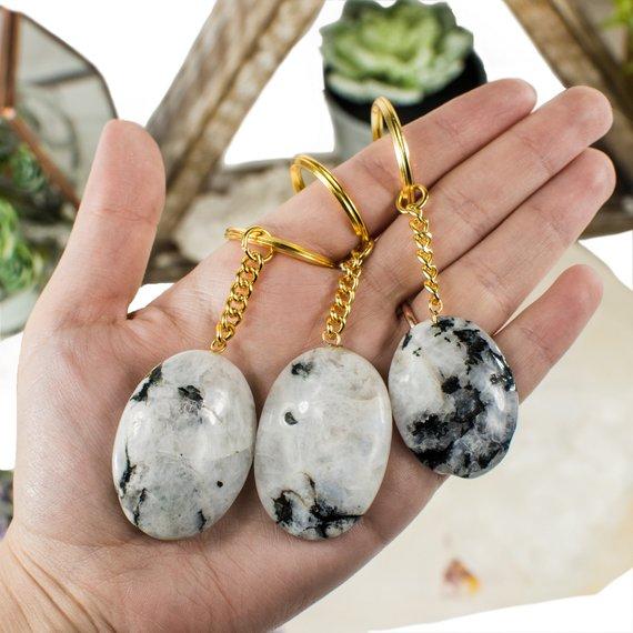 Hand holding up 3 Rainbow Moonstone Worry stone Keychains with various decorations blurred in the background