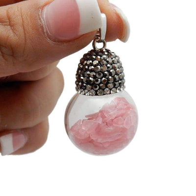 One silver plated glass ball pendant filled with rose quartz chips and a rhinestone top dangling from a woman's fingers.