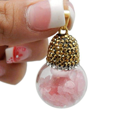One gold plated glass ball pendant filled with rose quartz chips and a rhinestone top dangling from a woman's fingers.