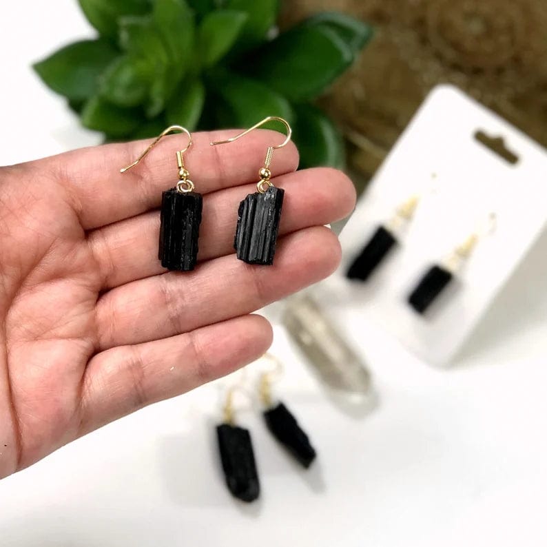 A pair of Black tourmaline rod earrings with gold ear wires in a woman's hand.  Two more pairs are blurred in the background.