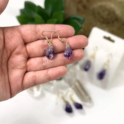 A pair of amethyst point earrings with gold ear wires in a woman's hand.  Two more pairs are blurred in the background.