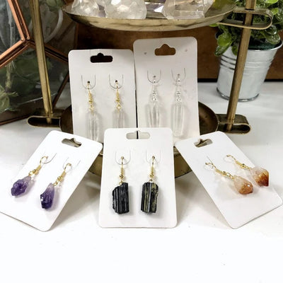 A photo of assorted crystal point earrings on white hang cards.  Pictured are crystal point earrings with gold ear wires, crystal point with silver ear wires, amethyst point with gold ear wires, black tourmaline with gold ear wires and citrine point earrings with gold earwires.