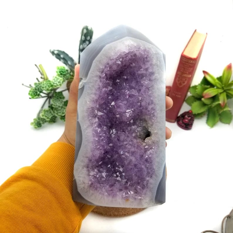 Amethyst cut base.  It has purple clusters in the center with white agate banding and a layer of gray on the outside. It is being held in a woman's hand.
