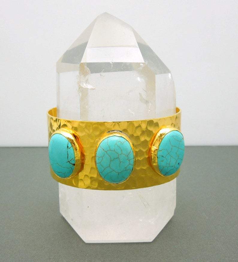 One gold plated cuff bracelet with three turquoise howlite stones on a gray background.  It is placed on a clear quartz crystal for display.