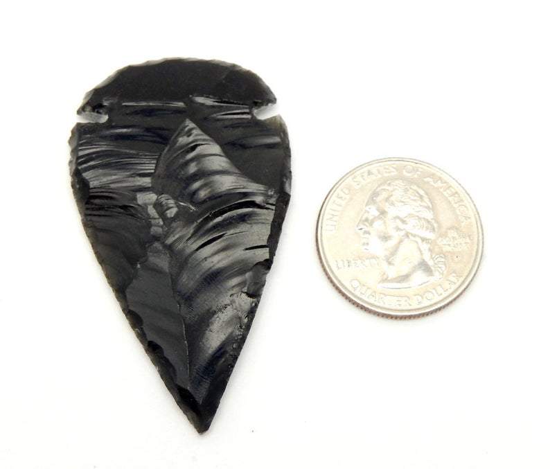 A black obsidian arrowhead next to a quarter on a white background showing it is about twice the size of the quarter.