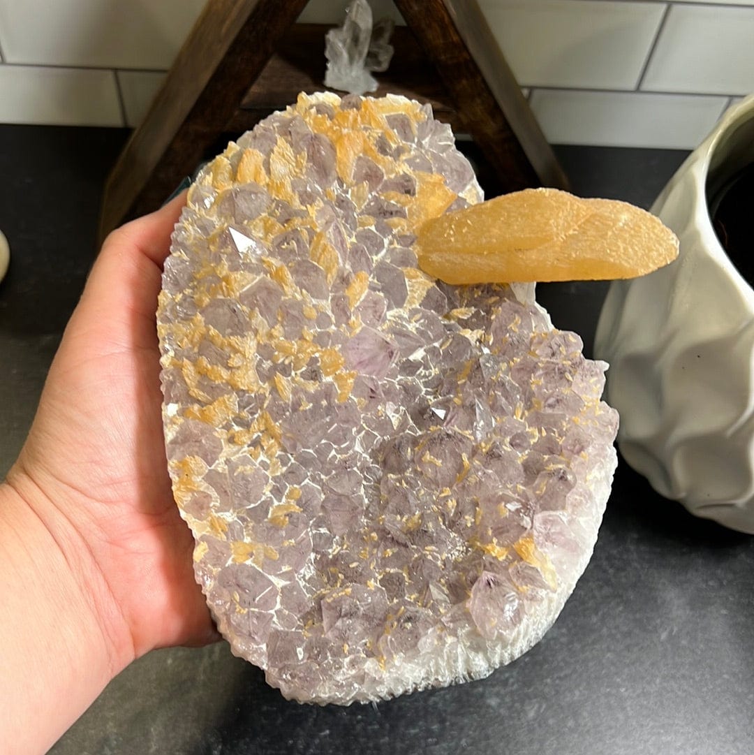 Amethyst cluster light purple with small orange calcite sprinkled along the top amethyst and one long orange calcite rod on the left side.