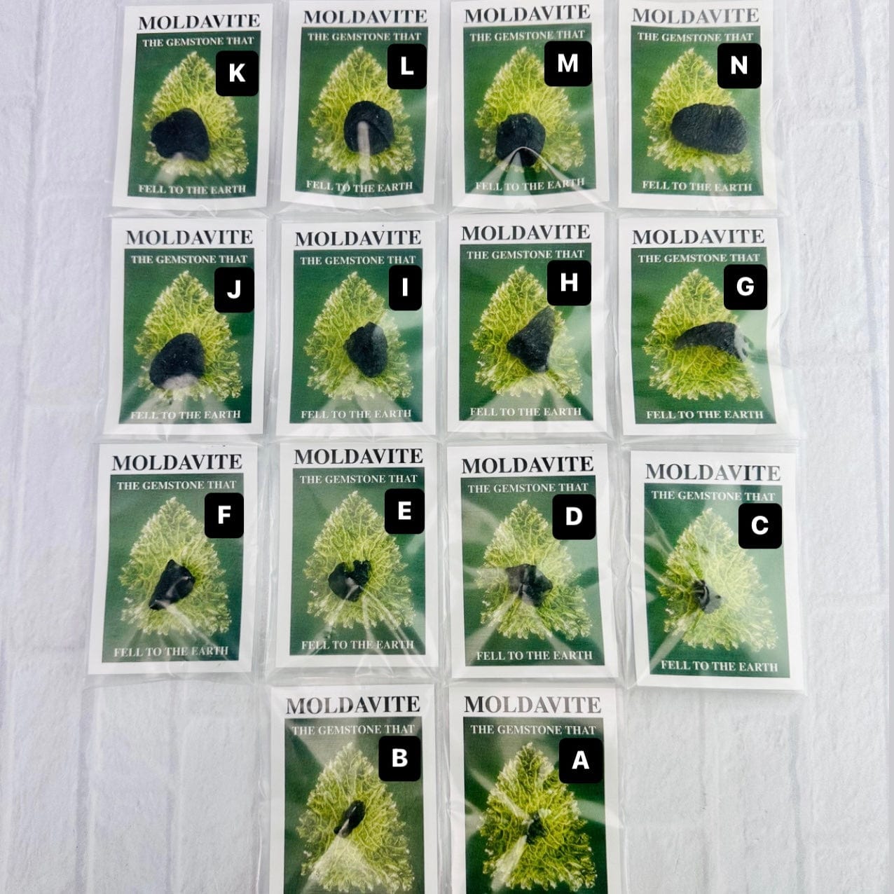 All fourteen Moldavite pieces displayed in their bags lined up on a white surface, with their respective letter next to each stone.