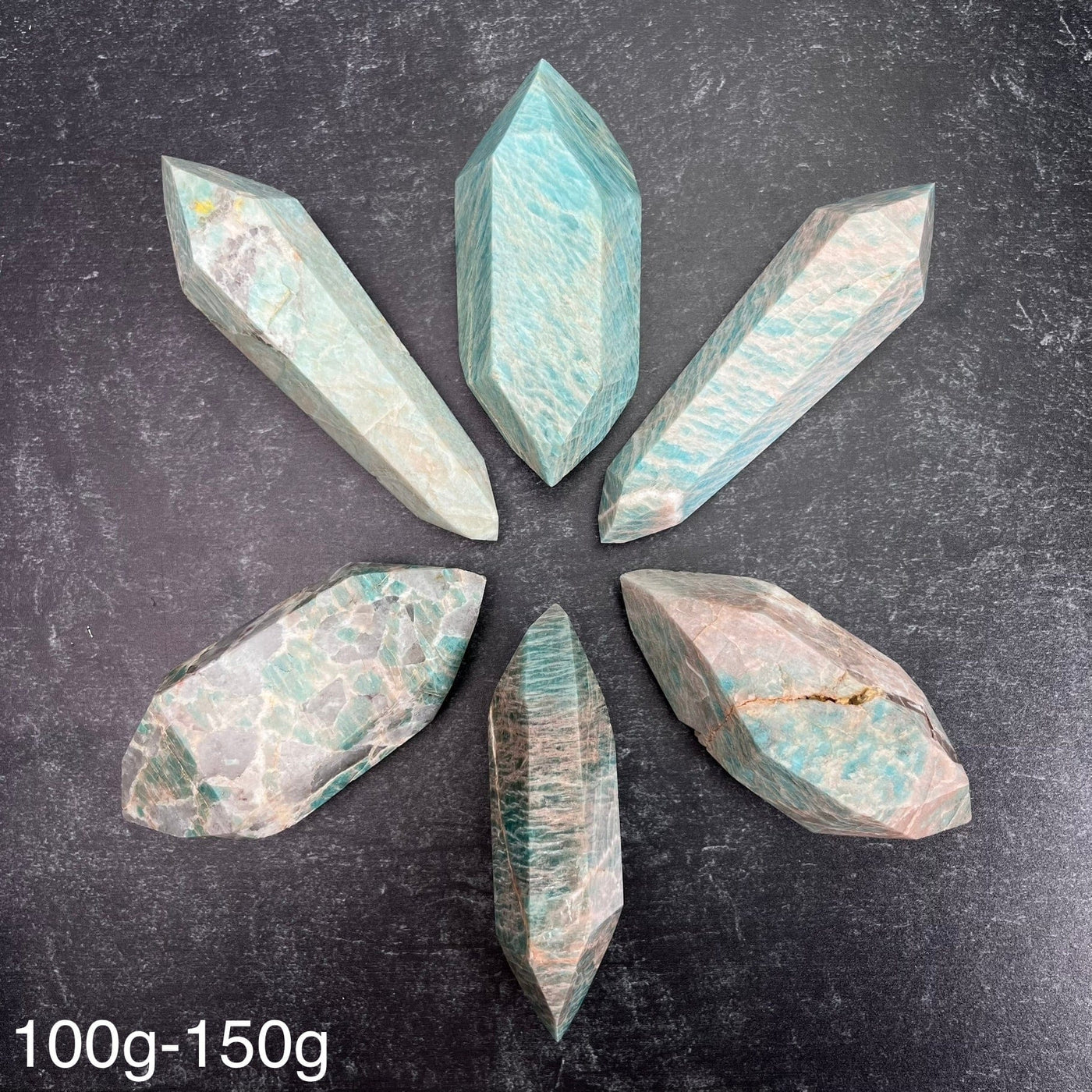 Six different variations of the Amazonite Polished Double Terminated Points, in the weight range of 100g-150g, laid out on a black surface.
