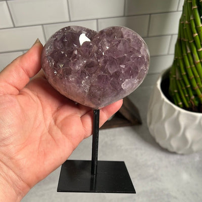 Amethyst cluster heart on a black metal stand.  Held in a hand.
