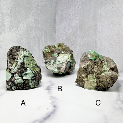All three stone choices (labeled A, B & C) of Rough Emerald Matrixes, on a marble surface.