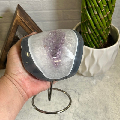 Agate polished stone with amethyst in the center on a silver metal base pictured on a gray background and held in a woman's hand.