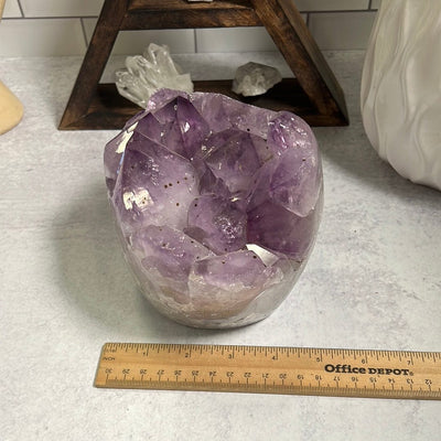 Amethyst cluster with polished sides.  The sides are more white and the top clusters are purple with some black speckles.  Next to a ruler showing it is around 4.5 inches wide