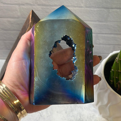 Agate polished point that has been titanium treated.  It is mostly blue with hints of rainbow colors created by the coating.  The center is open with druzy growing inside.  Held in a hand.