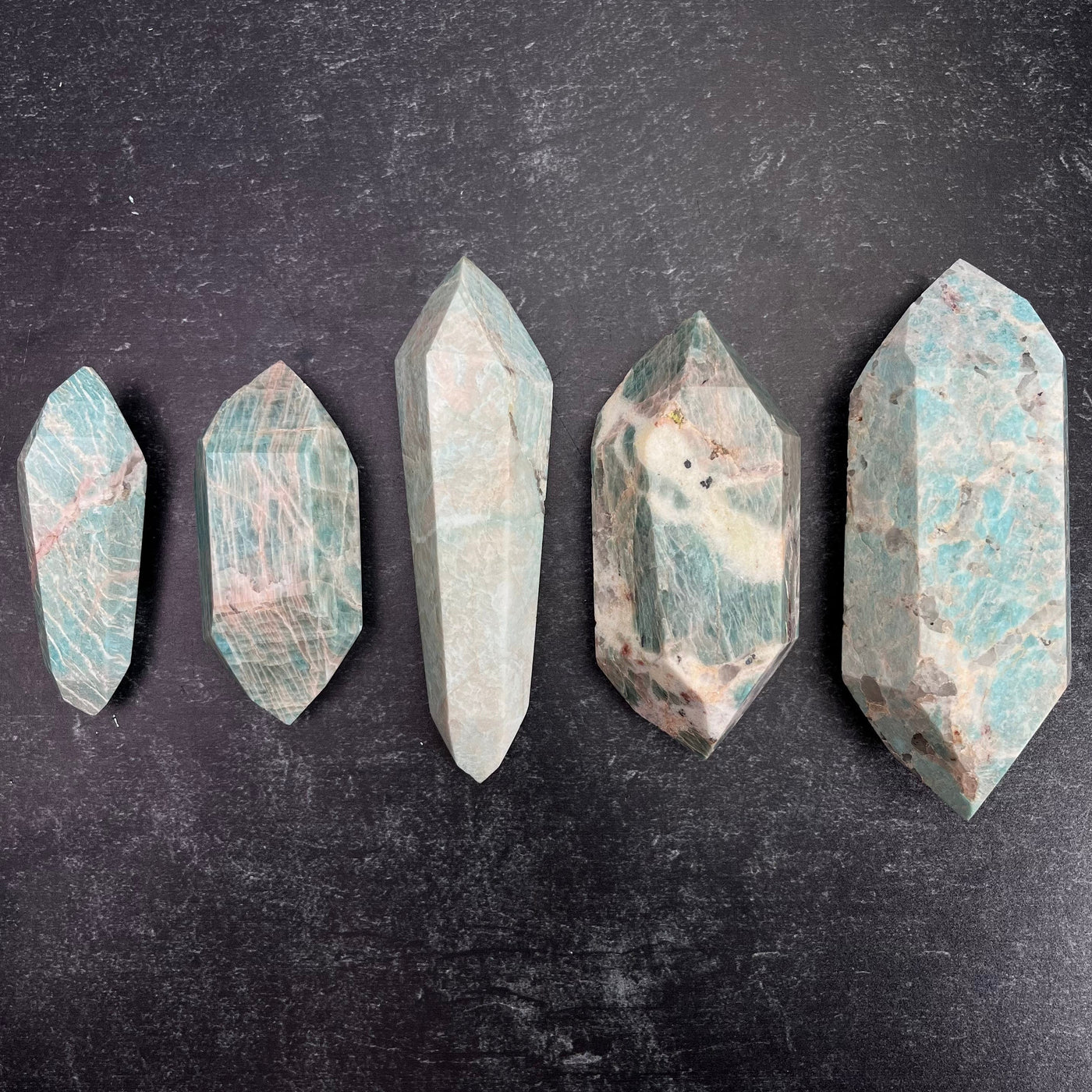 Five different weight options available of the Amazonite Polished Double Terminated Points. One of each displayed from smallest to largest, left to right on a black surface.