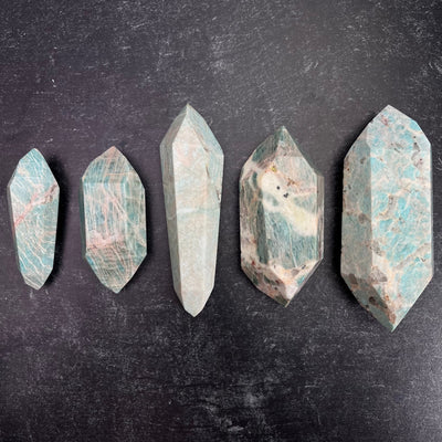 Five different weight options available of the Amazonite Polished Double Terminated Points. One of each displayed from smallest to largest, left to right on a black surface.