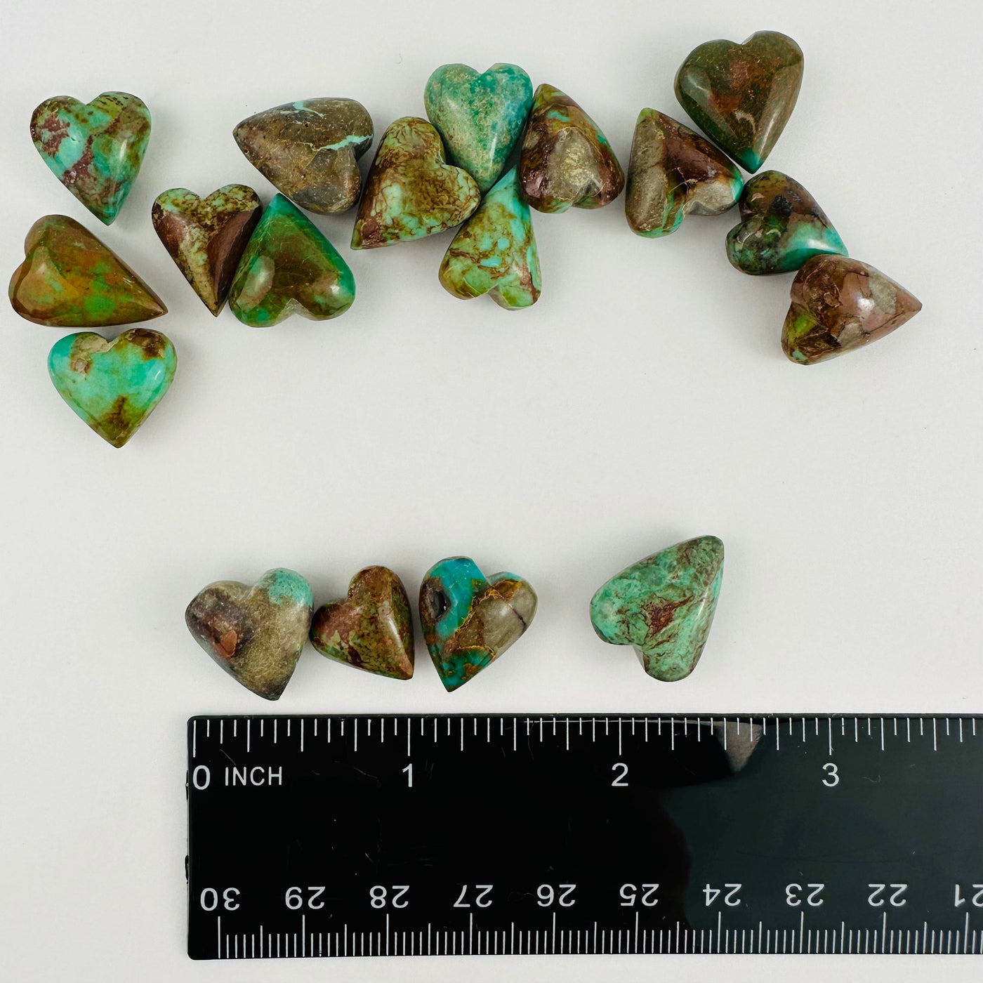 Turquoise hearts on a white background with a few by a ruler showing they are about half an inch but vary.