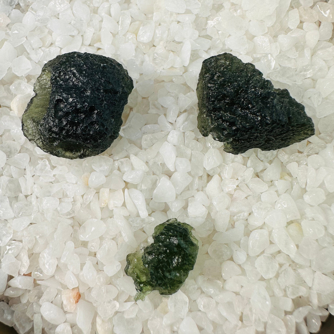 Three different sized Moldavite pieces displayed on top of crystal quartz chips.