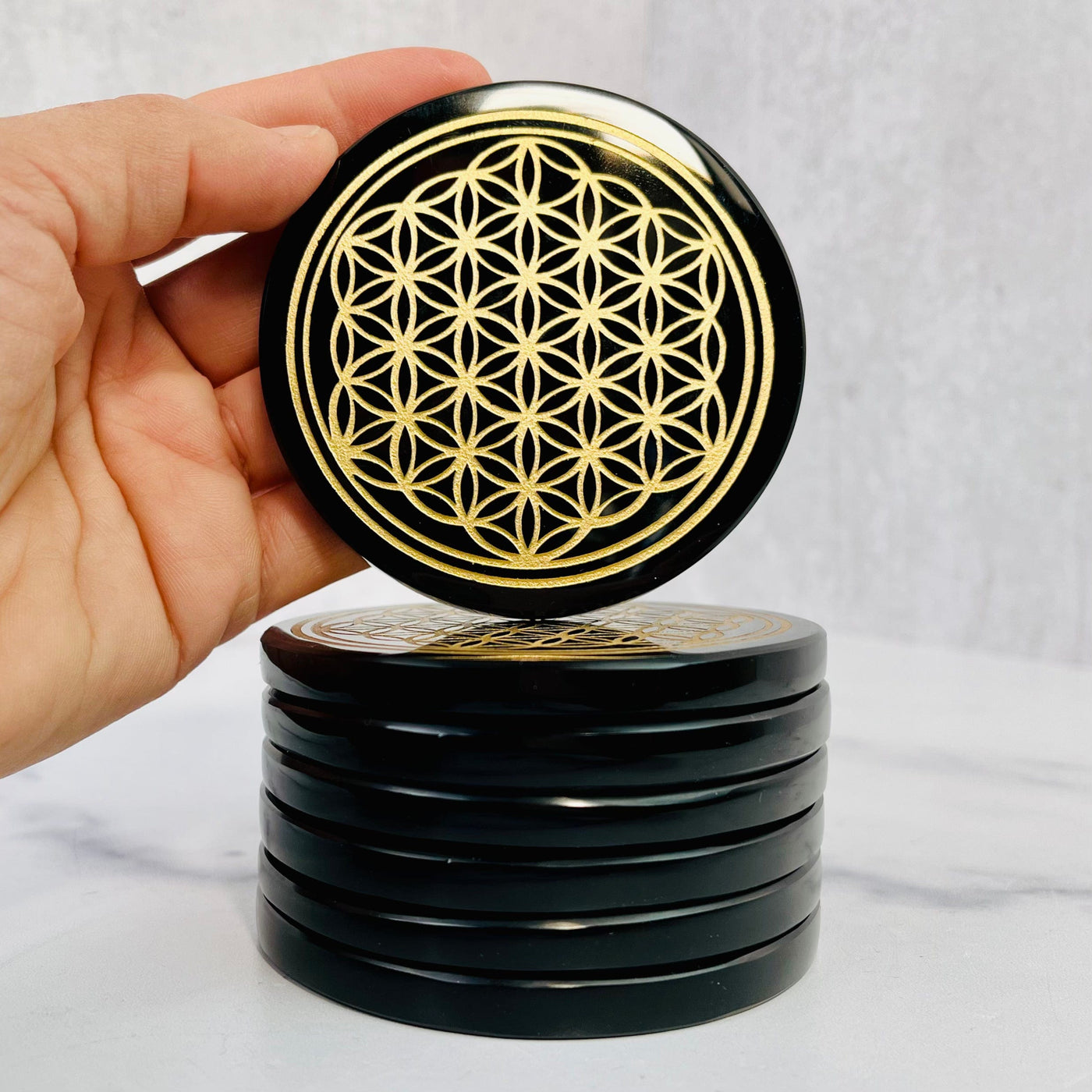 Stacked Black Obsidian Coasters with top one held up by woman's hand to show the flower of life engraving.