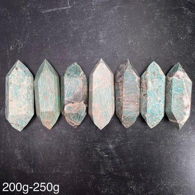 Seven different variations of the Amazonite Polished Double Terminated Points, in the weight range of 200g-250g, lined up on a black surface.