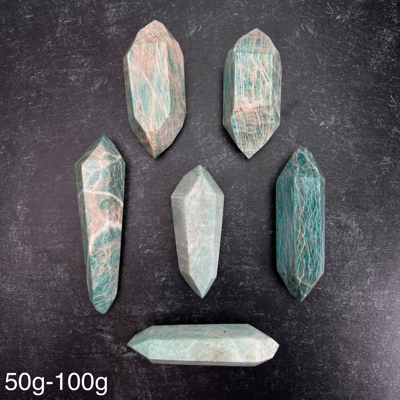 Six different variations of the Amazonite Polished Double Terminated Points, in the weight range of 50g-100g, laid out on a black surface.