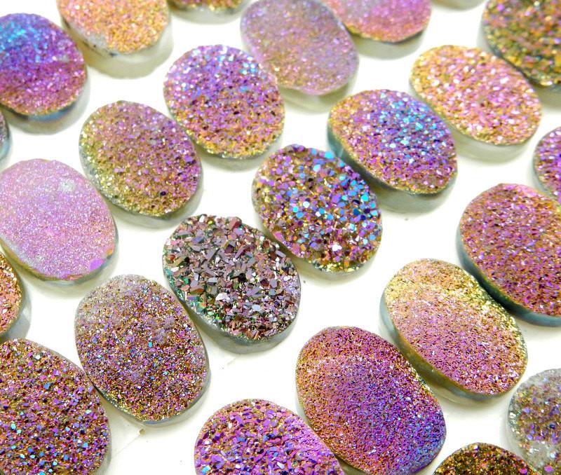 Up close view of various Purple And Yellow Oval Druzy Cabochons lined up on a white surface.