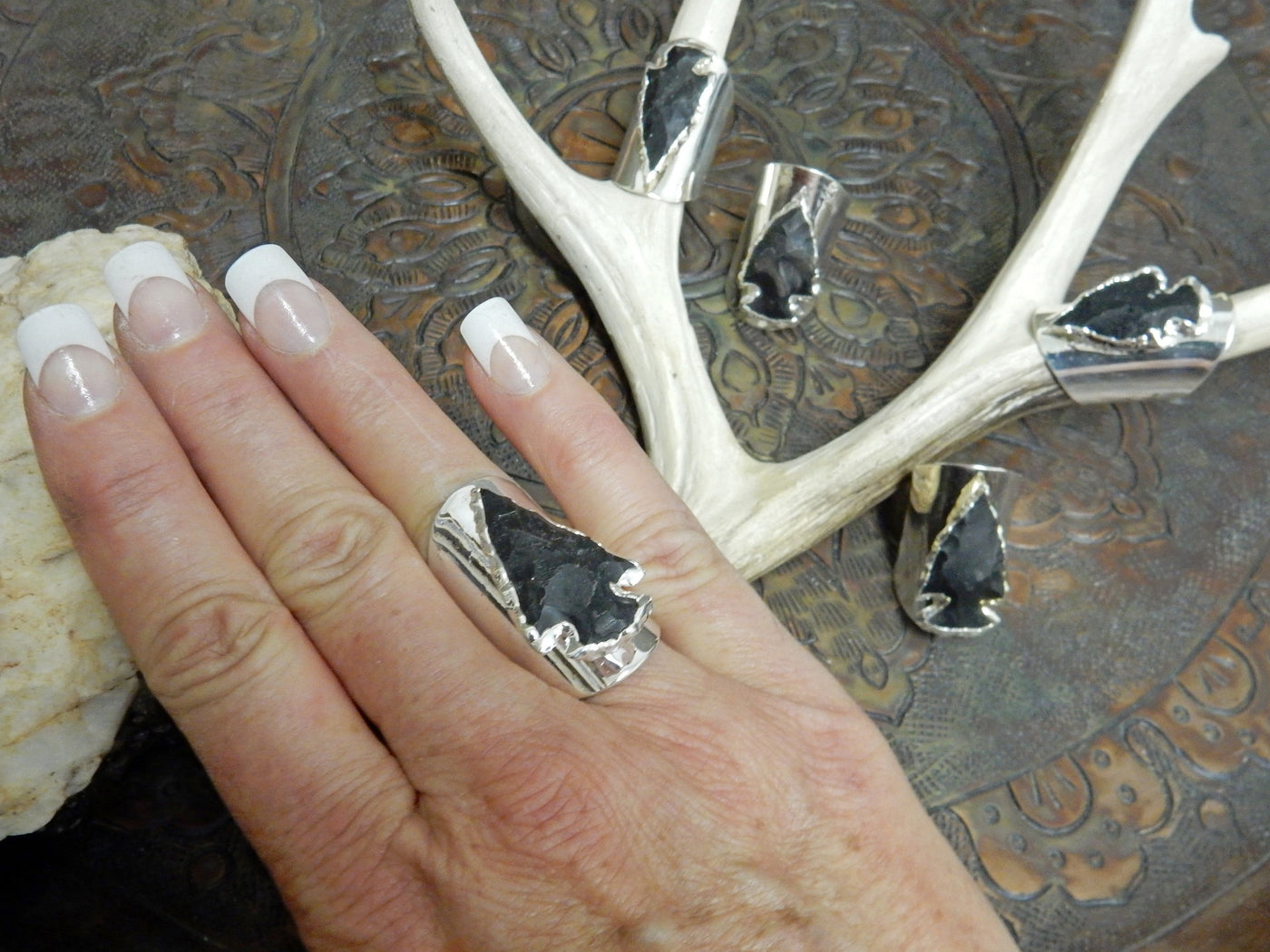 finger wearing Black Obsidian Silver edged Arrowhead set on Adjustable Silver Electroplated Cigar Band Ring with others in the background