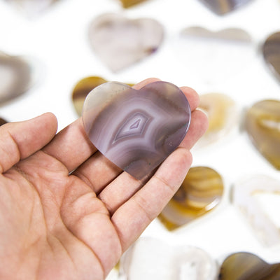 An Agate Heart Shaped Cabochon in a hand, multiple agate hearts in the background on a white surface.
