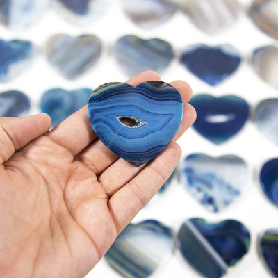 A blue Agate Heart Shaped Cabochon in a hand, multiple agate hearts in the background on a white surface.