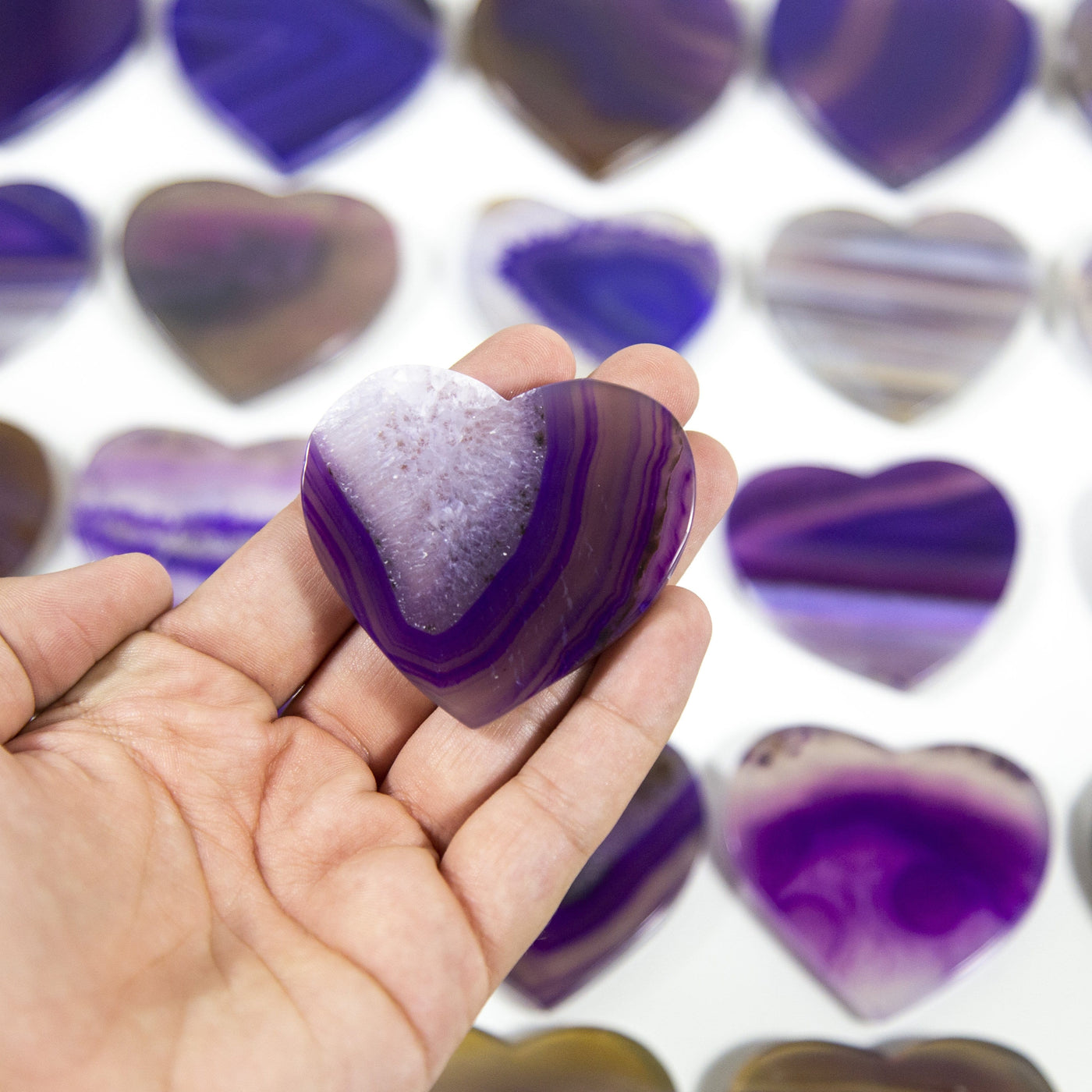 A purple Agate Heart Shaped Cabochon in a hand, multiple agate hearts in the background on a white surface.