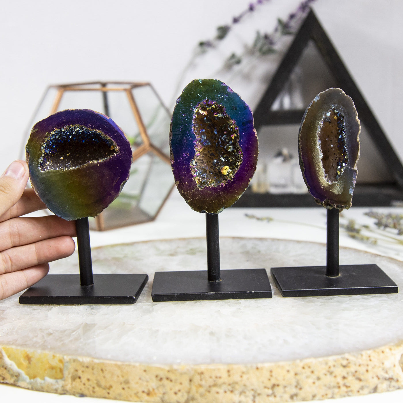 3 Rainbow Aura Geodes on Stands with a hand holding one on the left 