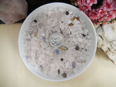 Rose quartz chips in a white bowl with a quarter in the bowl.  They are tiny and many can fit in the quarter.