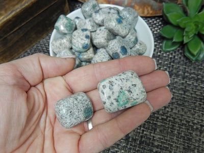 hand holding up 2 granite/jasper tumbled stones with others in the background