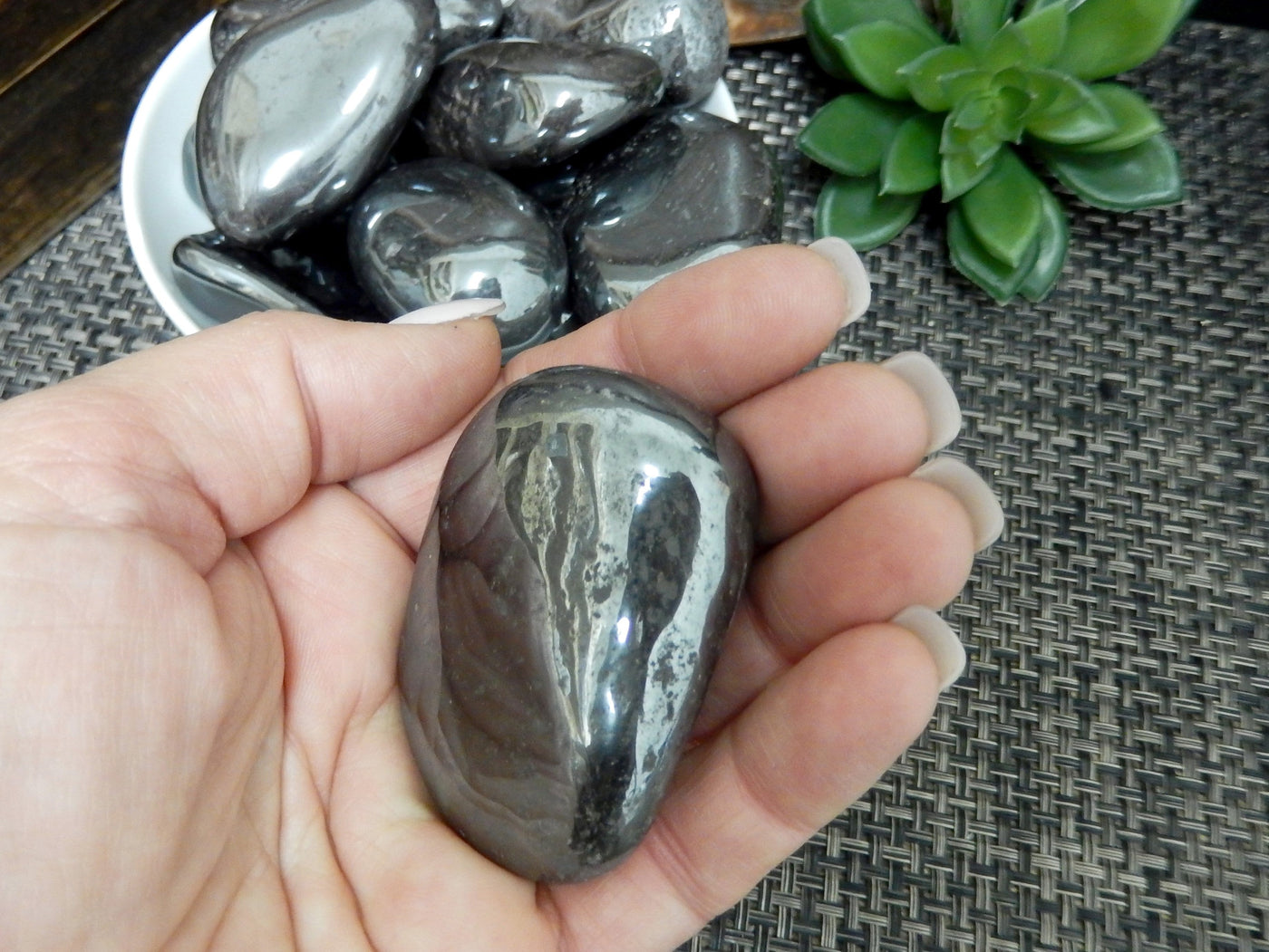 hand holding up hematite tumbled stone with others in a bowl in the background